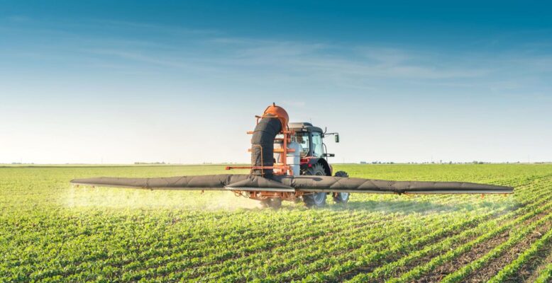 Das Bild zeigt einen Traktor, der über ein großes landwirtschaftliches Feld fährt und Pestizide versprüht. Diese Szene verdeutlicht den Einsatz von Pflanzenschutzmitteln in der Landwirtschaft, was zur Problematik von Pestiziden im Trinkwasser beitragen kann. 