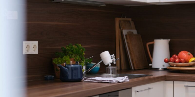 Das Bild zeigt eine moderne Küche mit einem Riva Alva S Jova Wasserhahnfilter der Firma riva, der am Wasserhahn installiert ist. Der Filter läuft und füllt ein Glas mit Wasser. Auf der Küchenarbeitsplatte stehen eine blaue Teekanne, frische Kräuter, eine Schüssel mit Tomaten und Kürbis sowie ein Wasserkocher.