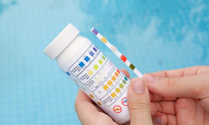 Nahaufnahme einer Hand, die einen Wasserteststreifen neben einem Testbehälter hält, um den pH-Wert mittels einer Farbindikation und andere Wasserparameter wie der Wasserhärte eines Pools zu messen. Der Behälter zeigt farbcodierte Referenzwerte zur Analyse von Chlor, pH und Alkalinität